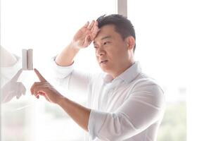 Asian businessman in office heat, chooses cooling mode of air conditioner on control panel on white wall photo