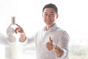 Portrait of a smiling Asian man. He stands by the wall of the building, presses the incoming call, control panel with his hand. He looks at the camera, shows a super. photo