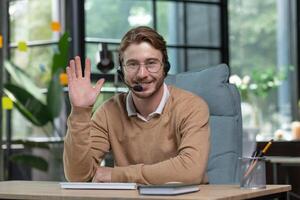 Online trade, support, consultation. Portrait of a young handsome man sitting in the office at the table in a headset, working at the computer, looking at the camera, smiling, waving his hand. photo