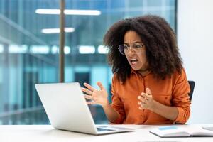Young businesswoman expressing frustration while looking at her laptop. She appears angry and stressed, gesturing with her hands. The modern office setting suggests a work-related issue. photo