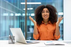 A frustrated office worker with hands raised in confusion, sitting at her desk with a laptop. The image captures a common workplace scenario of stress and uncertainty. photo