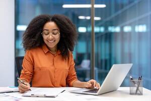 A young businesswoman smiles while working at her desk, using a laptop and writing notes. Her friendly demeanor and focused attitude convey professionalism and positivity in a modern office setting. photo