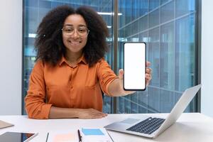 A cheerful woman in an office setting shows a smartphone with a blank white screen. The modern work environment includes a laptop, notebook, and window view. photo