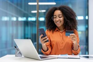 Young businesswoman using phone and credit card, smiling confidently at her desk. The setting implies a modern office environment with technology and finance playing key roles in her workflow. photo