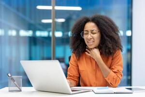 An office worker holds her throat in discomfort while sitting at a desk with a laptop. She appears to be dealing with a health issue during work hours, looking visibly uncomfortable. photo