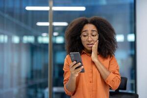 A young businesswoman appears shocked while checking something on her phone in a modern office setting. Her expression suggests receiving unexpected news or encountering a surprising situation. photo
