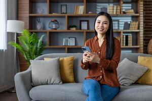 Asian woman using smartphone while sitting on couch in modern living room. Smiling, relaxed, casual atmosphere, enjoying home comfort while engaging with phone technology. photo