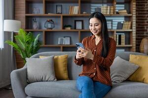 Asian woman sits on sofa using smartphone, smiling while engaged in online activity. Modern home setting with stylish decor and comfortable seating creates relaxing environment. Technology in everyday life. photo