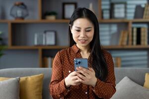 Asian woman casually dressed, sitting on couch at home, using smartphone. Smiling and engaged in activity, creating warm and relaxing domestic atmosphere. Perfect for lifestyle concepts. photo