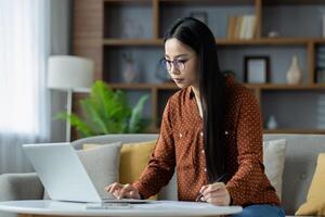 Asian woman working from home on laptop, deeply focused in stylish living room. She uses pen and paper for notes, showcasing remote work lifestyle and concentration in cozy environment. photo