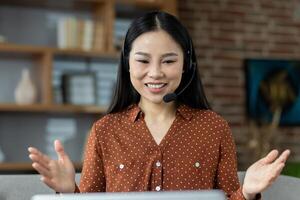 Asian woman in a virtual meeting, wearing headset, smiling and gesturing. Shows remote work and online communication in comfortable home setting. Captures professional yet relaxed atmosphere. photo