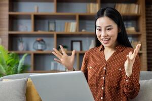 Asian woman using laptop at home, appearing joyful and surprised. Engaged in call, showcasing expressive emotions and connection. Casual attire enhances comfortable home setting. photo