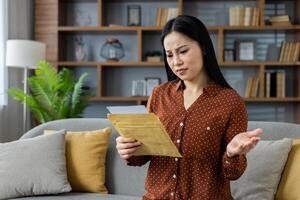 Asian woman at home reading a letter with confused expression on sofa. She appears thoughtful and concerned, reacting to unexpected news in a modern living room setting. photo