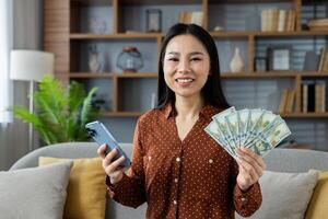 Asian woman holding smartphone and showing dollar bills, smiling confidently in home setting. Represents financial success, digital transactions, modern lifestyle. Phone indicates technology usage. photo