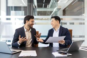 Businessmen conversing in office while reviewing documents, showcasing teamwork and collaboration. Smiling professionals fostering communication and productivity. Laptops and notes on table. photo