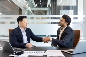 Businessmen shaking hands in modern office. Image captures partnership, trust, successful deal. Laptops, charts, documents on desk highlight professional environment. Concept of teamwork, cooperation photo