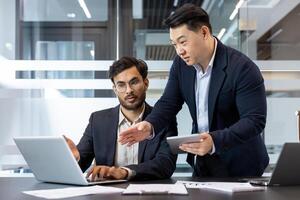 Two business professionals engage in a collaborative discussion in a modern office environment. One stands holding a tablet, while the other works on a laptop. The setting highlights teamwork. photo