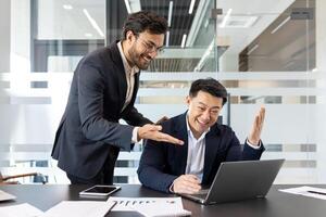 Two businessmen are joyfully celebrating a success in a modern office setting. One is using a laptop, while the other gestures excitedly. Their expressions show happiness and teamwork. photo