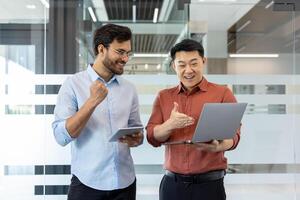 Two happy colleagues celebrate successful project in modern office. One holds laptop, the other a tablet, showcasing teamwork, achievement, and excitement in business collaboration with technology. photo