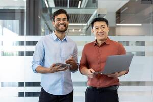 Two smiling businessmen stand in a modern office environment holding a tablet and laptop. Their casual attire complements the professional setting, indicating a collaborative atmosphere. photo