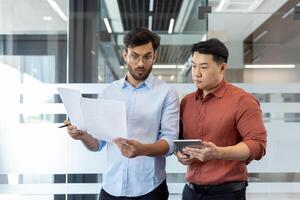 Two male professionals engage in a discussion over paperwork in a modern office setting. They appear focused and engaged, emphasizing teamwork and communication in a corporate environment. photo