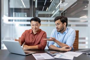 Two colleagues engaged in a lively discussion at a modern office table with laptops and papers, highlighting teamwork and collaboration. The office setting is sleek and contemporary. photo