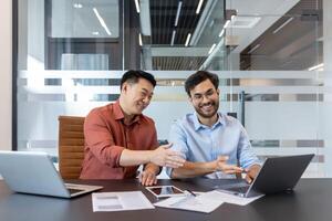 Two male colleagues are engaging in a discussion while working on laptops in contemporary office setting. Papers and smartphone are also visible on the desk, highlighting a collaborative environment. photo