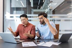 Two frustrated businessmen sit at office table with laptops, papers, and phone, discussing challenges during online meeting, showing expressions of disbelief and concern. photo