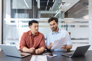 Two professionals engage in a discussion over important documents in office. Laptops and a phone are on the table, suggesting a tech-driven work environment. Collaboration and focus are evident. photo