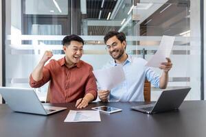Business colleagues celebrate success with laptops and documents. Smiling coworkers review project results on paper and phone, highlighting teamwork and achievement in modern office. photo