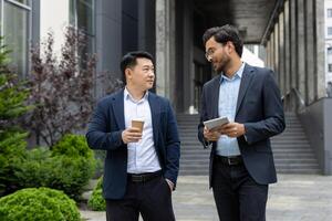 Two professional men in business attire engage in a friendly discussion while holding coffee cups and tablet. The setting is urban, with greenery and modern architecture in the background. photo
