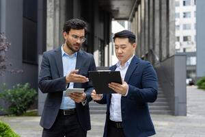 Businessmen discussing work in corporate environment. One holding tablet, another with clipboard, focused on conversation. Modern setting enhances business strategy context. photo