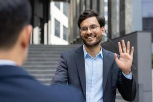Businessman in suit greeting colleague with friendly smile and wave. Outdoor meeting in city, showcasing professional interaction and positive connection during workday. photo