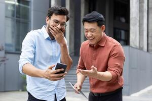 Two men share a joyful moment, laughing while looking at something on a smartphone. They appear to be outside an office building, enjoying a break together. photo