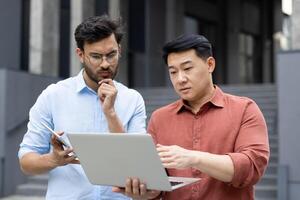 Two focused businessmen collaborate outdoors, using laptop and tablet for discussion. They appear engaged problem solving, bringing together modern technology and teamwork in a professional setting. photo