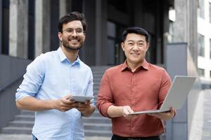 Two professionals, equipped with a tablet and laptop, are smiling while working outdoors. This scene suggests a collaborative business environment with a focus on technology and teamwork. photo