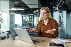 Mature business woman displays frustration during online conversation in office setting. Laptop and open notebook accentuate professional vibe, emphasizing challenges of remote communication. photo