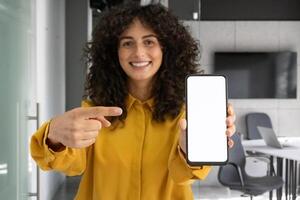 Smiling woman in yellow shirt points to blank smartphone screen in office. Concept of technology, presentation, business, communication, and digital display. Ideal for tech and marketing. photo