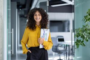 Confident businesswoma holding clipboard in modern office with plants and open laptop, presenting professional atmosphere. Ideal for themes of success, leadership, business. photo