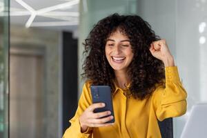 Excited woman celebrating success using smartphone in modern office. Happy expression, closed fist signifies achievement. Image portrays joy, communication, connectivity, and optimism with use phone. photo