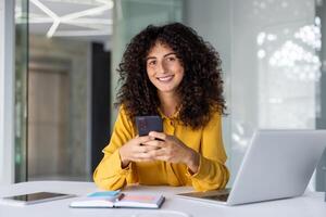 Smiling woman holds smartphone in modern office setting with laptop and notebook. Represents work, technology, communication, success, and confidence. Ideal for business and tech themes. photo