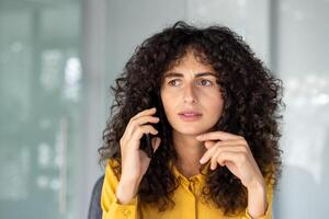 Curly-haired woman in yellow shirt using smartphone, appears concerned during phone call in office. Thoughtful and focused expression suggests work-related matters. photo
