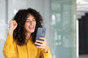 Joyful woman in yellow shirt celebrates success with smartphone in modern office setting. Displaying cheerful expression and energetic gesture while using phone. Highlighting happiness achievement. photo