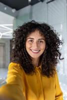 Smiling woman taking selfie using phone indoors. Curly hair and joyful expression highlight modern, bright setting. Perfect for concepts of happiness and connectivity. photo