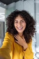 A cheerful woman takes a smartphone selfie in an open-office environment. Her friendly smile and outgoing energy are perfect for illustrating concepts like happiness and connection. photo
