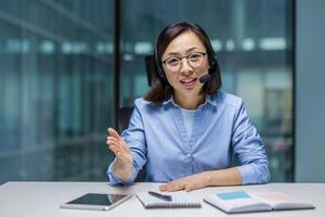 An Asian businesswoman using a headset to communicate during a virtual meeting. She is seated with documents and a tablet on the table. Ideal for concepts like communication photo