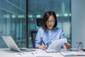 Asian woman concentrates on analyzing data at her office desk. She reviews documents while using a laptop, exemplifying professionalism and focus in a modern workspace. photo