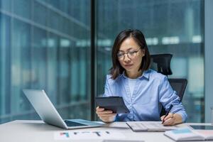 An Asian woman is deeply engaged in accounting work at a modern office desk. She is using a tablet and laptop to manage financial tasks efficiently. Her serious expression reflects concentration photo