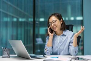 Asian businesswoman on phone call in office. Smiling and confident while using laptop, showcasing modern work environment and communication. Captures professionalism and productivity. photo