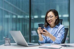 An Asian businesswoman in a modern office setting using phone. She is smiling and appears engaged with her work, sitting at a desk with a laptop and notebook. photo